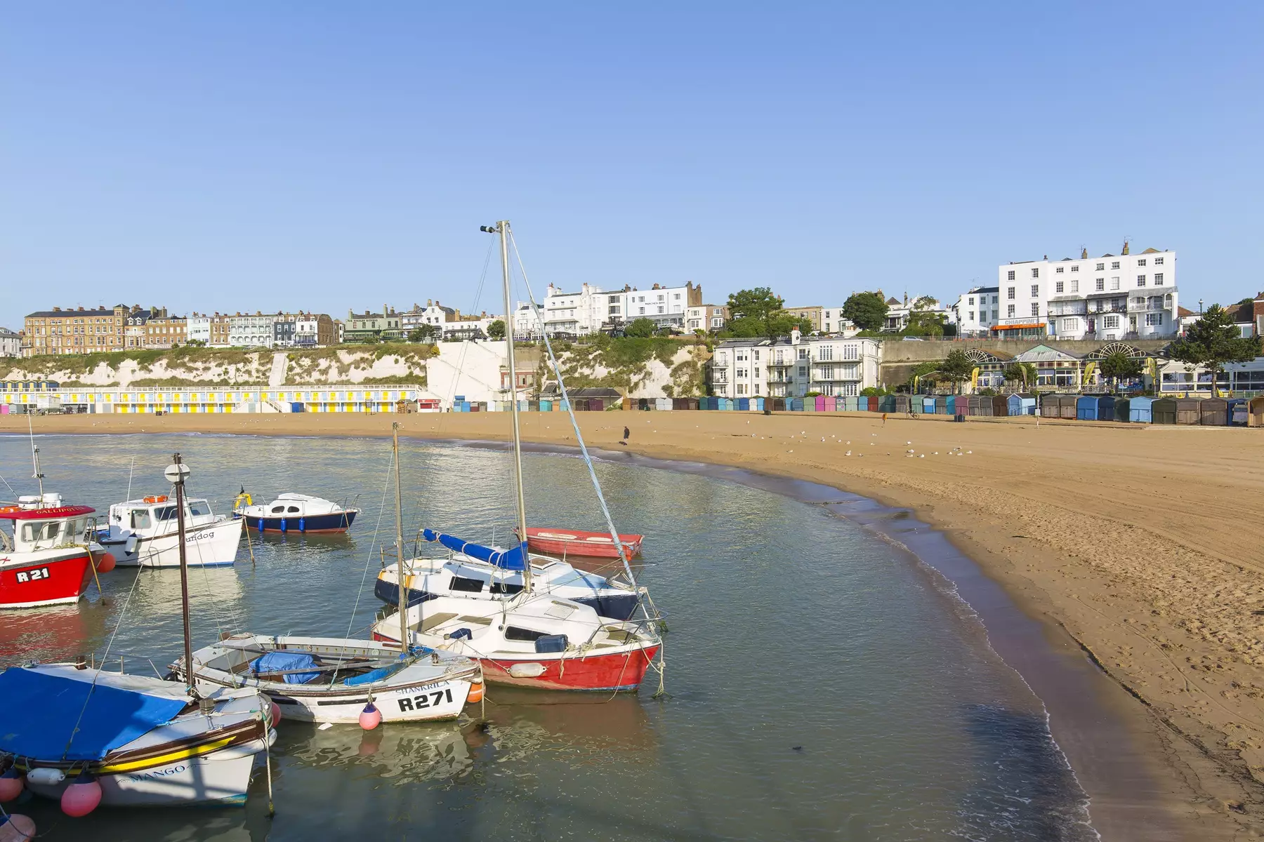 Boats in the harbour close to the beach with cliffs and building lining the back of the beach